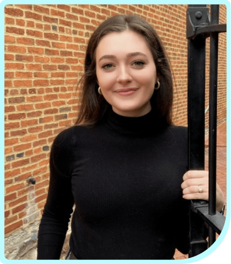 A young woman in a black turtleneck shirt standing in front of a brick wall.